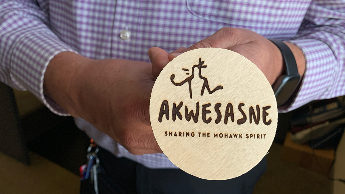 A close up of a man's hands holding a wooden circle with a laser engraving that reads "Akwesasne Sharing the Mohawk Spirit"