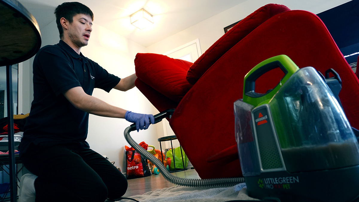 Gavin Decontie kneels down and uses a small carpet cleaner on a red sofa