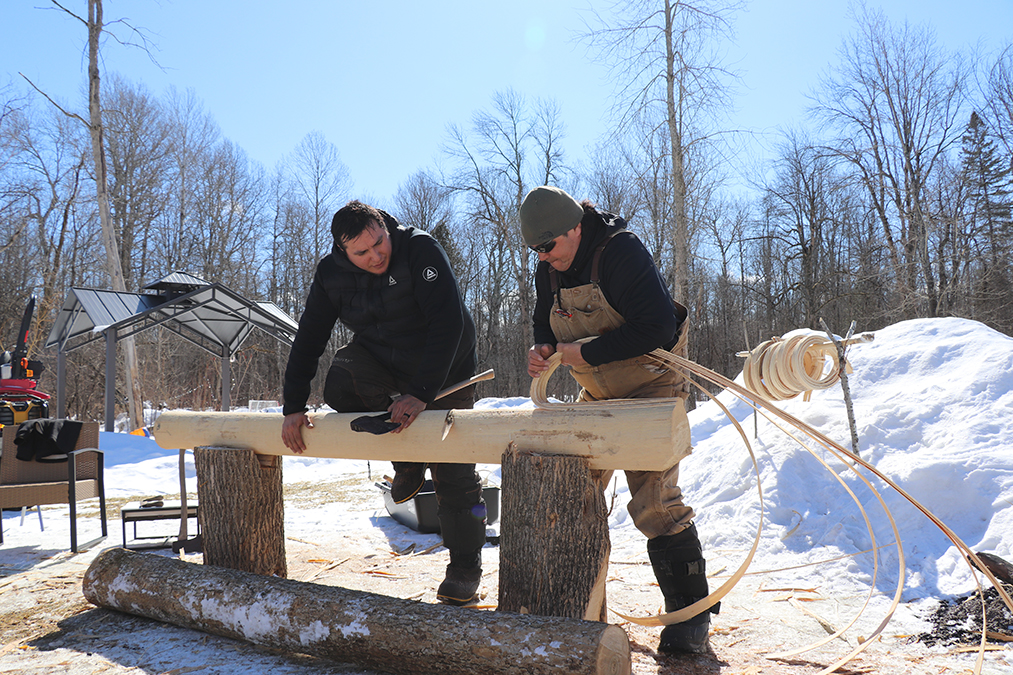 feature photo Two men stand next to one another, outside on a sunny day, hunched over a log. The man on the right pulls long strips of wood off the log as the man on the left watches, leaning in with his axe in his hand.
