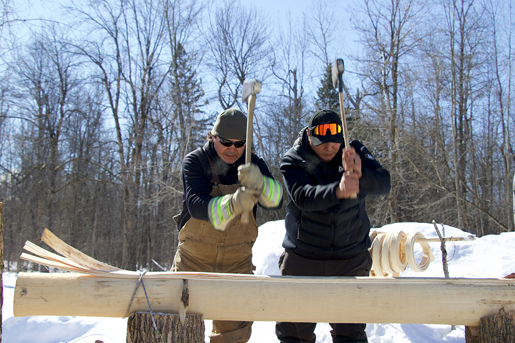 photo 2 Angello Johnson and Eric Sunday outside on a sunny day pounding a black ash log with the back end of an axe.