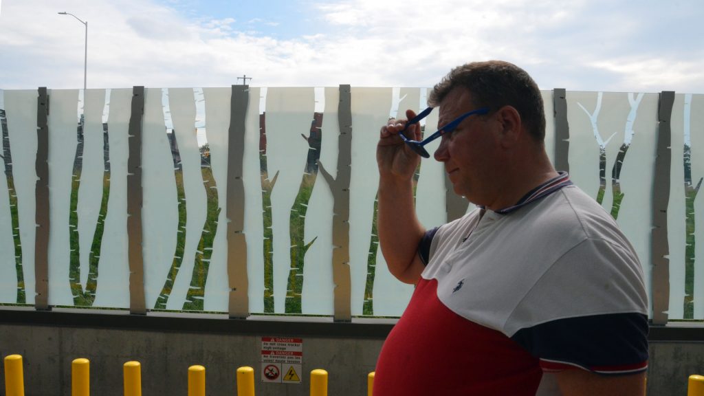 A man walks in front of a glass and metal fence with a row of birch trees making up the exterior fence at Pimisi station.