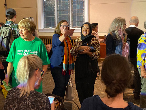 NDP candidate Emilie Taman is shown speaking with an audience member in the crowd.