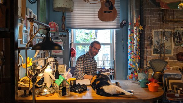 A man sits at the front desk of a cluttered store, smiling as he pets a cat that lays on the counter.