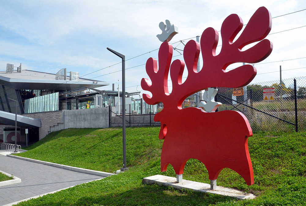 A red moose statue stand in the grass behind Pimisi station.