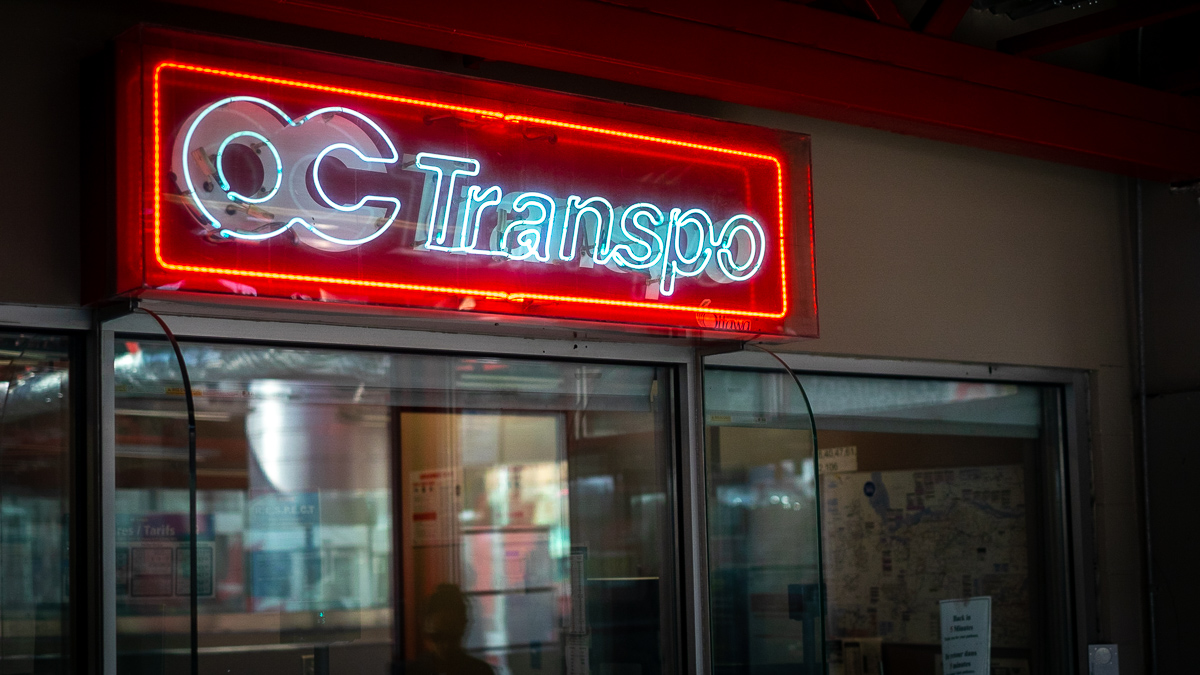 A neon OC Transpo sign hangs above a booth at an LRT station.