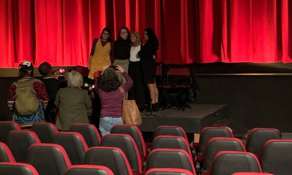 4 young girls stand on a stage in front of a red curtain posing for pictures while a few people take their picture in front of movie theatre seating.