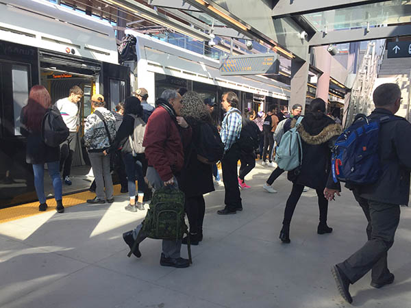 Riders coming and going from a stationary train, the sun shines down on the station. 