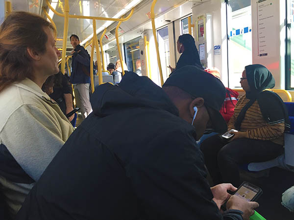 Passengers inside the train, some are seated others standing.  The sun shines in through the window on the far side.