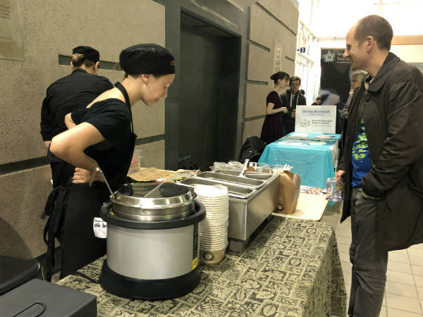 Woman standing at a table serves hot vegan meals to a man during the film festival.