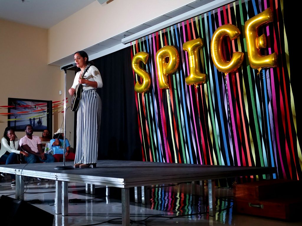 Young woman plays guitar and sings into microphone on stage.