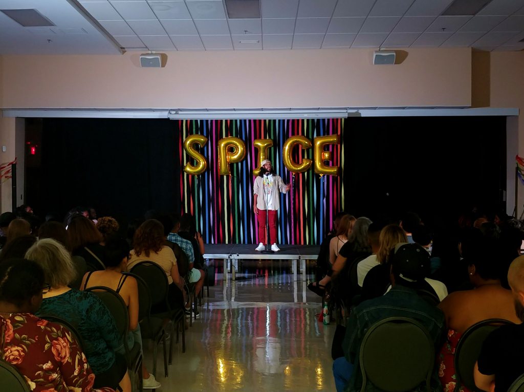 Young man speaks into microphone on stage and gestures towards the crowd.