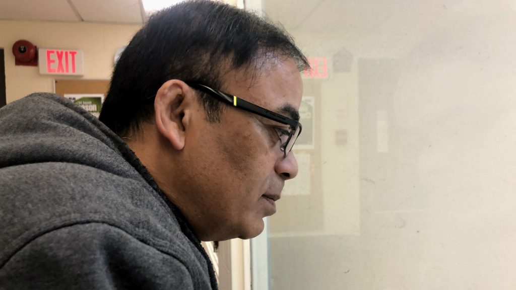 Man with glasses looks down at the squash courts through a window in an upstairs viewing area.