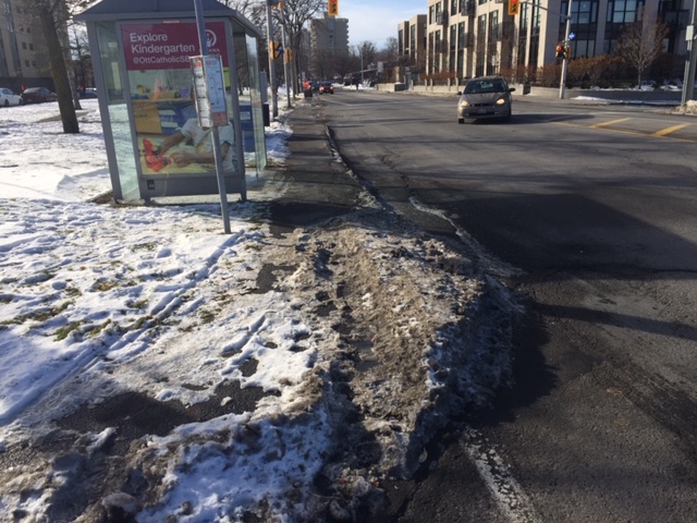A snowy sidewalk near a bus stop, with muddy footprints on the surface creating a barrier.