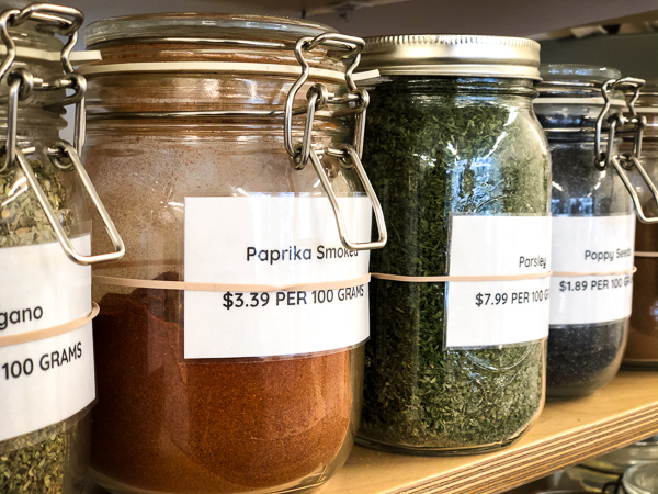 A close photo of 4 jars of herbs and spices sitting on a shelf: oregano, paprika, parsley and poppy seeds