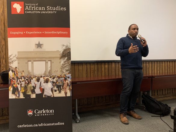 Councillor Rawlson King speaking with a microphone beside a tall banner for the Carleton University Institute for African Studies at an Afro-Caribbean Mentorship Program event at Carleton University on Friday, November 29.