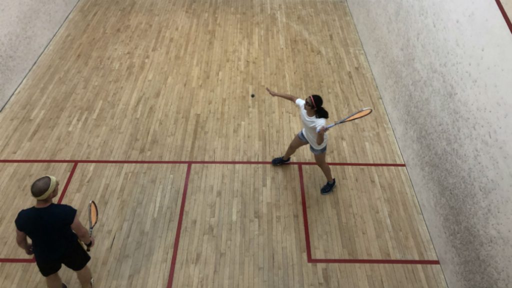 Squash player in white t-shirt and grey shorts captured mid-swing. Her opponent stands back, waiting to return the hit.