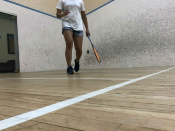 athlete walks to the service box, marked by white lines on the court. She bounces the ball and holds her racket at her side.