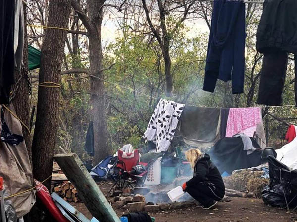 Woman crouches beside fire, clothes hang on lines in the surrounding trees at the Bayview 'tent city'