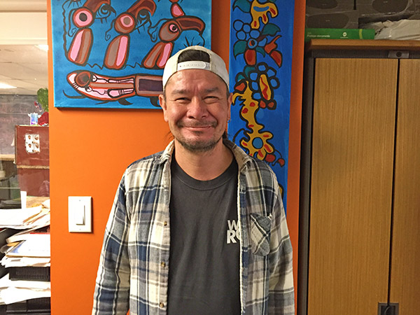 Windsor stands in front of an orange wall, smiling.  He wears a white backward hat and navy and white flannel.