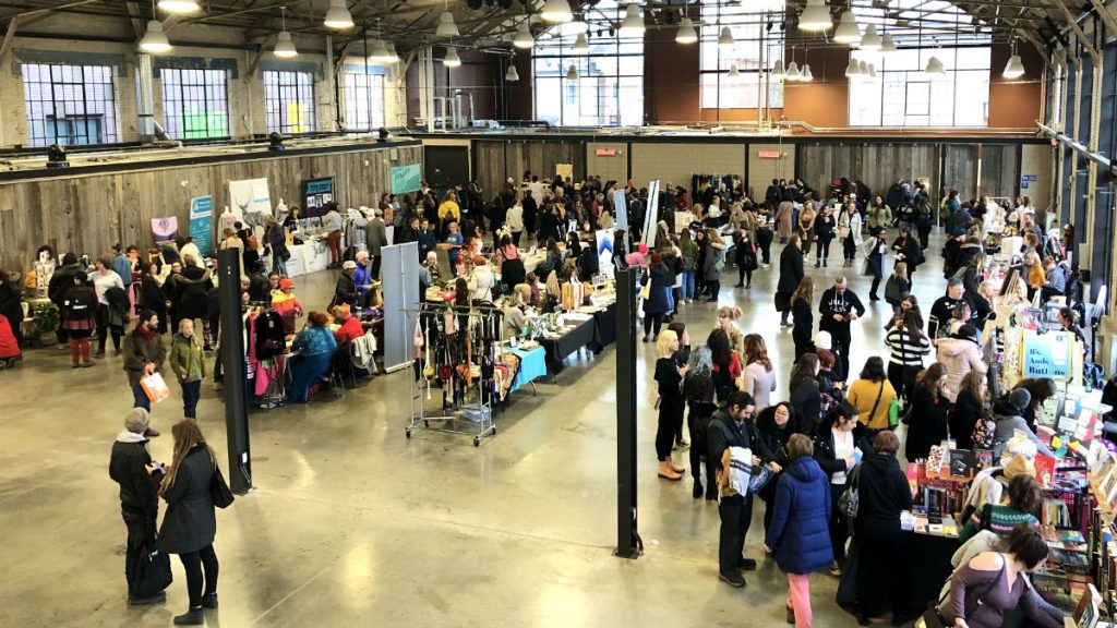 A crowd of around 100 people inside a large building browsing booths showcasing feminist-themed crafts and women support groups.