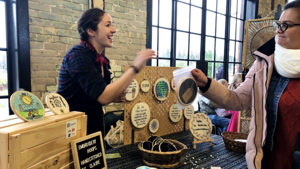 Claire Depoe-Collins, left, smiles and interacts with a customer while handing her a homemade cross-stitched sign from her vendor booth.