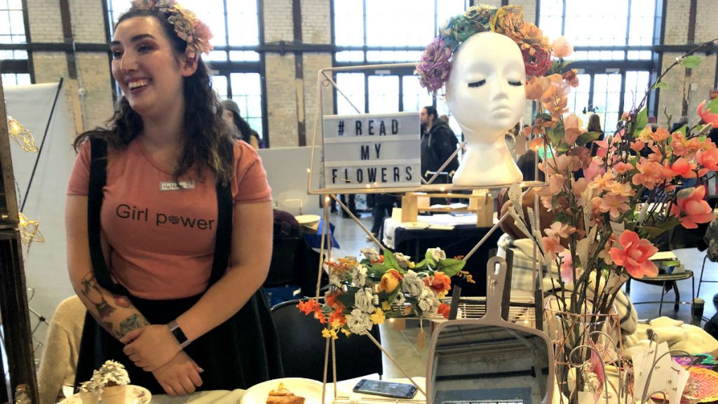 Helena Verdier smiles in a pink "Girl Power" t-shirt with flowers in her hair while conversing with a customer at her booth. She is surrounded by colourful flowers. A sign next to her reads," #ReadMyFlowers", the name of her business. 