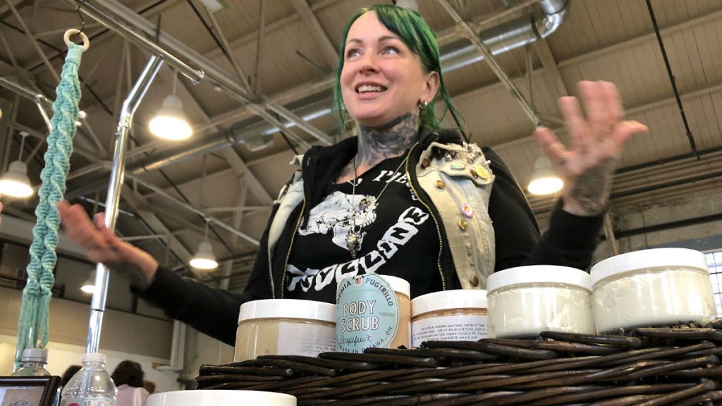 Danielle Montgomery has green hair, tattoos, and a black t-shirt. She stands behind her booth while holding her palms face up, explaining what feminism means to her.