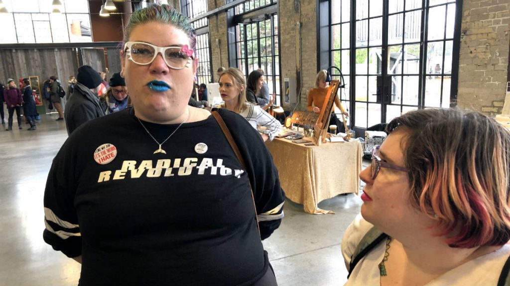 Ray Eskritt, left, wears bright blue lipstick, white glasses, and a shirt saying "Radical Revolution". Gabi Antaya, looks at Eskritt as they have a conversation about feminism. 