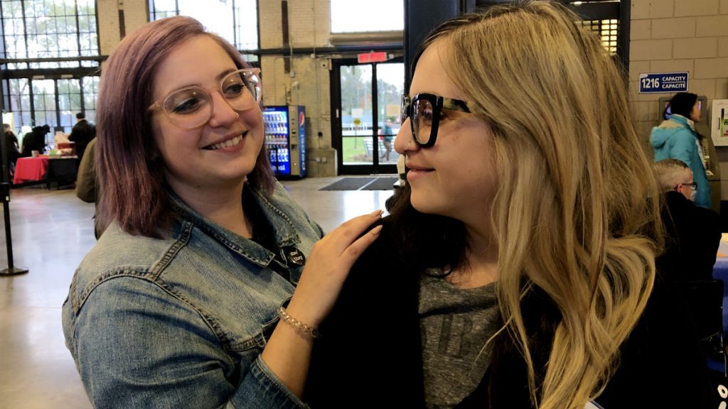 The Feminist Twins, Kayla and Jenna Spagnoli, smile at each other during the Feminist Fair. Kayla, with short purple hair and glasses, stands slightly behind her sister with her hand on Jenna's shoulder. Jenna wears glasses and has two-toned long blonde and black hair. 