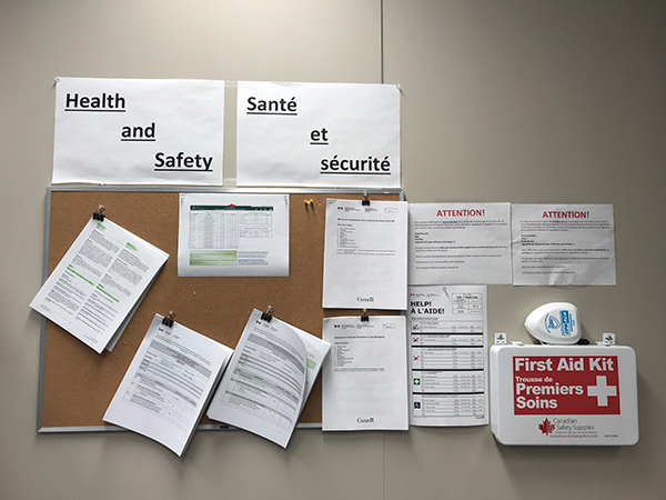 A bulletin board containing numerous documents surrounding health and safety protocols at the LEL building. To the right of it is a first aid kit. 