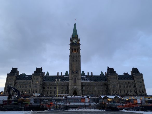 The Centre Block of Parliament, with the Peace Tower at the centre, and construction and fencing in the foreground. The clouds above are dark and grey.
