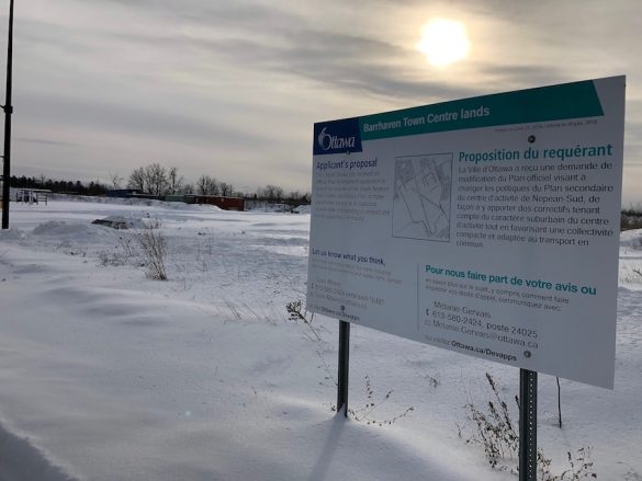 The business building proposal sign for Barrhaven's downtown core located in front of the undeveloped land that is currently covered in snow.