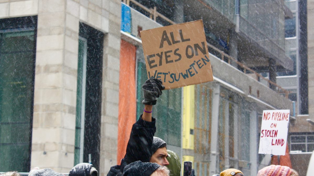 A hand in the middled of a crown holds up a cardboard, hand-written sign that reads "all eyes on wet'suwet'en"