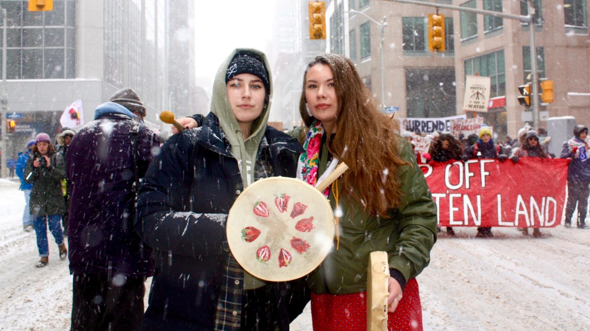 Two Indigneous women holding drums look at the camera, posed with the rally in the backdrop. 