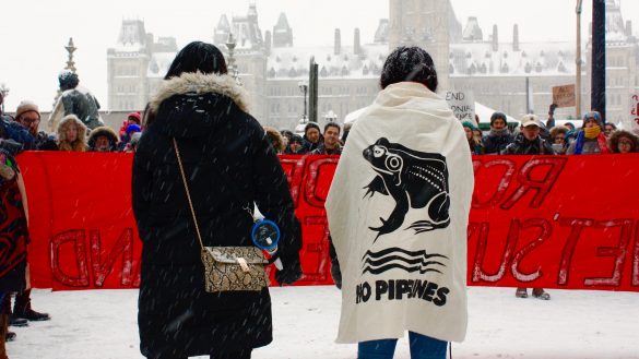 Photo is taken from behind. Two women stand in front of a crowd, behinda bright red banner that says "RCMP off Wet'suwet'en land". The girl on the right wears a canvas cape that says "no pipelines" with a frog on it.