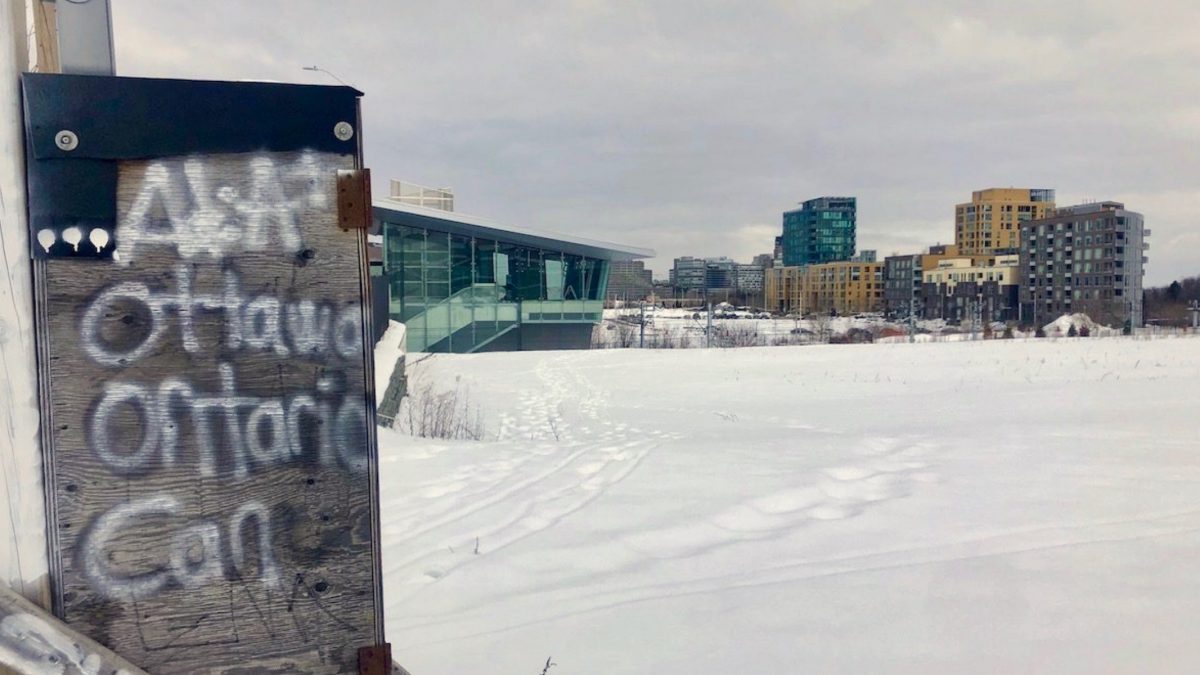 Large field covered in snow with Pimisi station and the Claridge Homes condo buildings in the background. Wooden box with "Ottawa, Ontario, Can" written on it in the foreground.