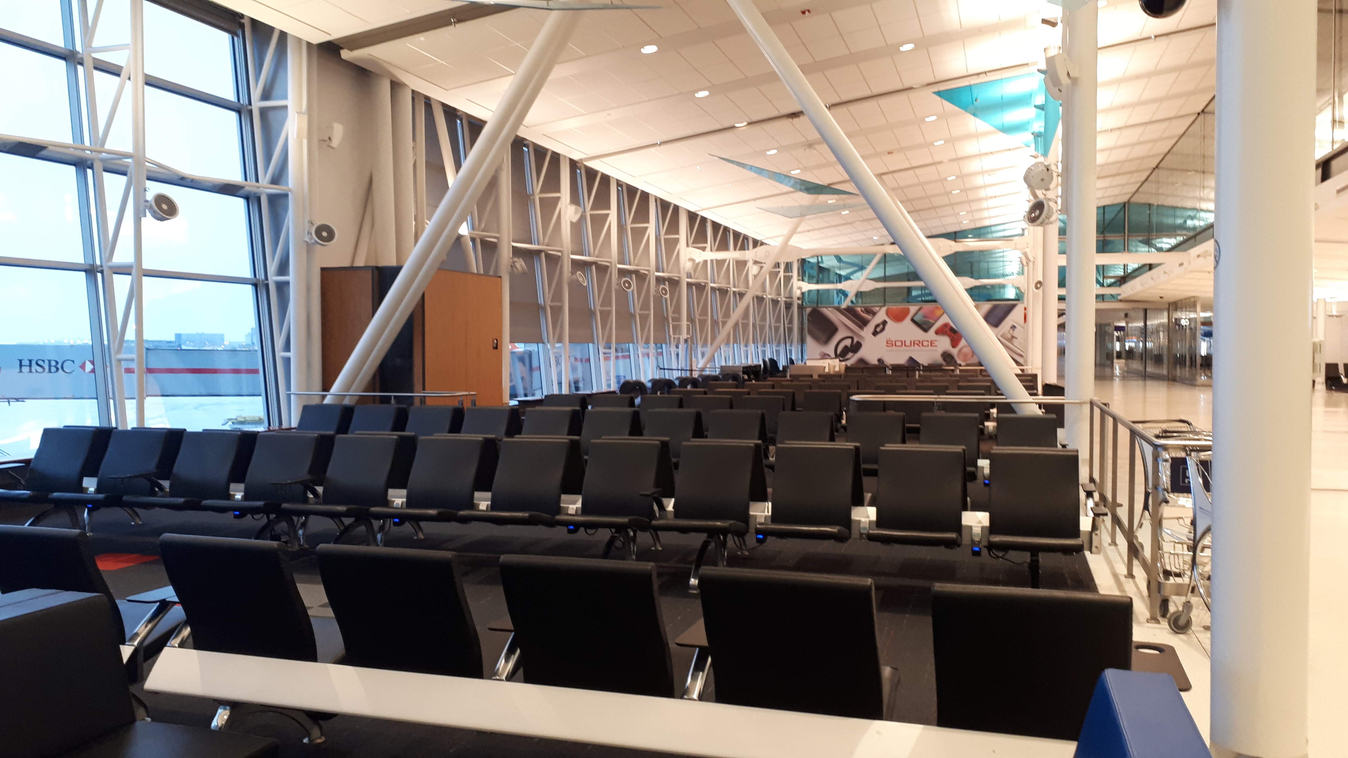 Several lows of empty seats. Large windows on the left reach up to the ceiling with blue morning light coming in. A long hallway can be seen on the right side of the image. Tall metal pillars are intterspersed in the image holding up a high ceiling. 