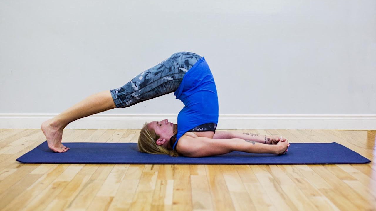 A woman in a blue shirt and workout leggings poses on a blue yoga matt with her legs over her head and arms stretched out. 
