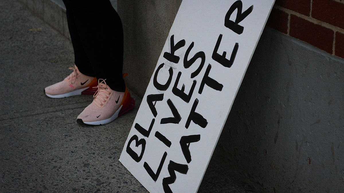 A sign that says "Black Lives Matter" leans against a brick wall as someone is pictured standing beside it.