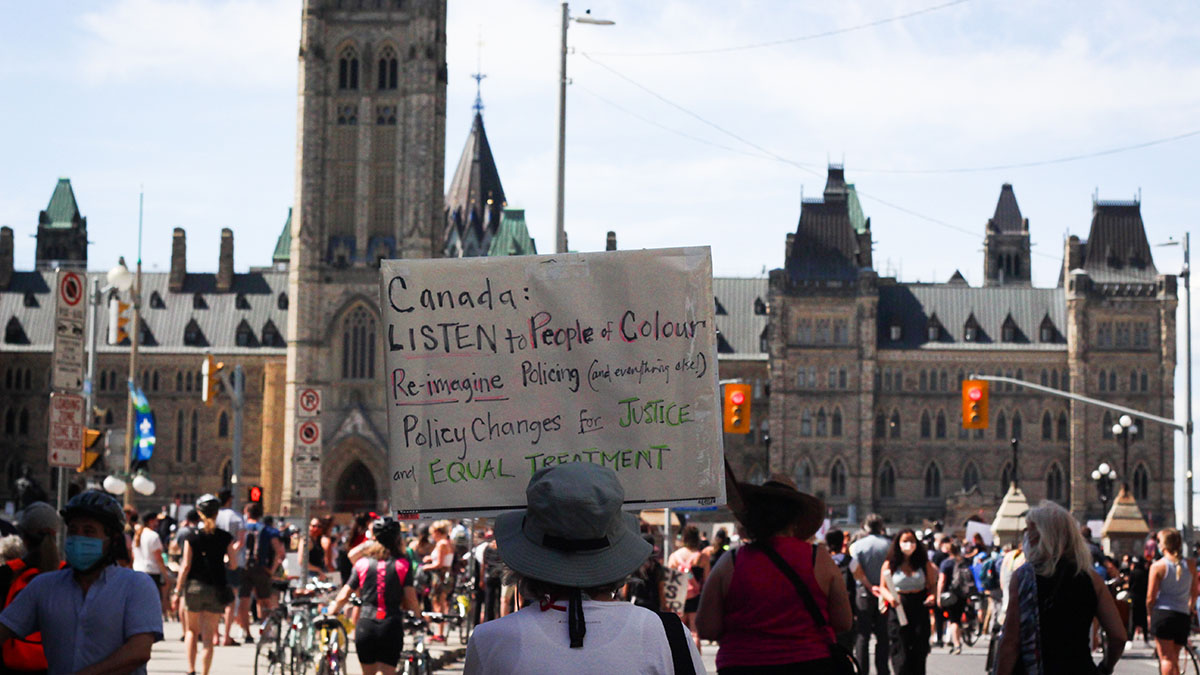 A protestor holds a sign that reads "Canada: listen to people of colour, re-imagine policing (and everything else!), policy changes for justice and equal treatment" in front of the parliament buildings