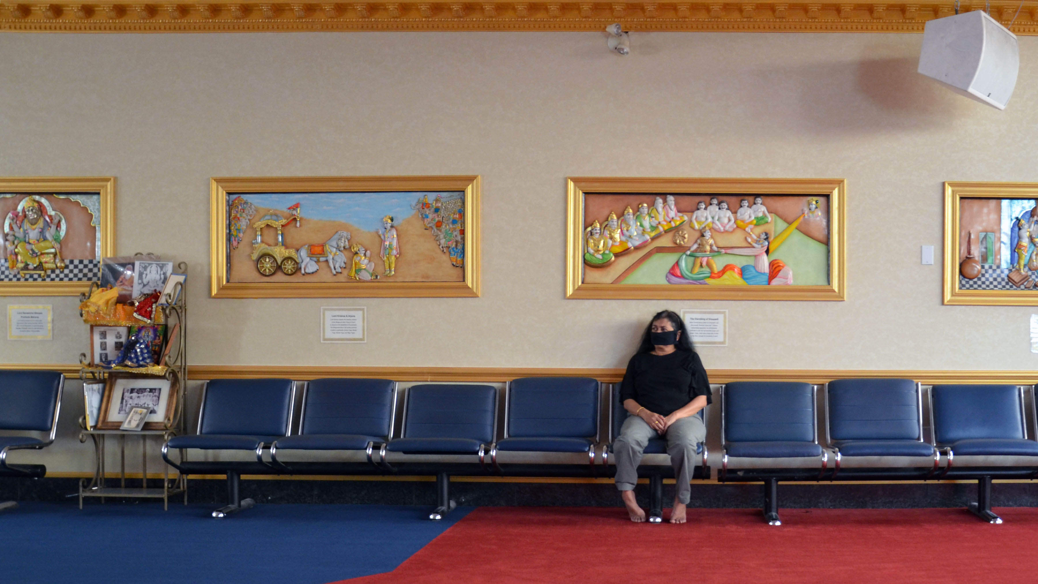 A masked woman sits alone on benches in a Hindu temple.