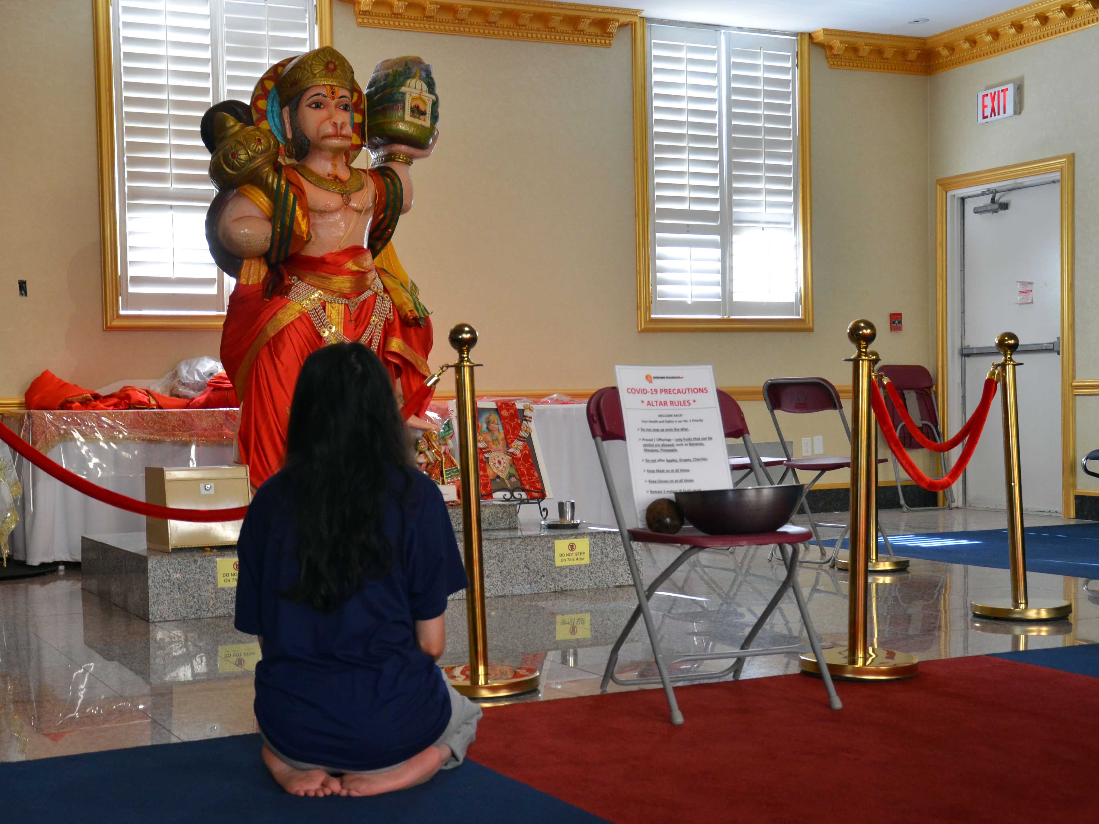 A woman kneels at an altar at Vishnu Mandir beside a sign detailing the temple's COVID-19 precautions such as "do not step up onto altar."