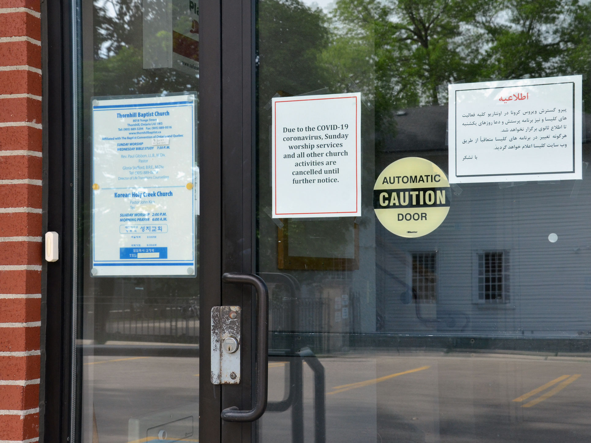 Amongst three signs on the front door of Thornhill Baptist Church, one  reads “due to COVID-19 coronavirus, Sunday services and all other church activities are cancelled until further notice.” An empty parking lot is reflected in the door. 