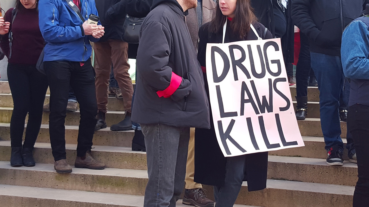 People stand on steps during a protest on the National Day of Action on the Overdose Crisis. The protest sign reads, "drug laws kill."