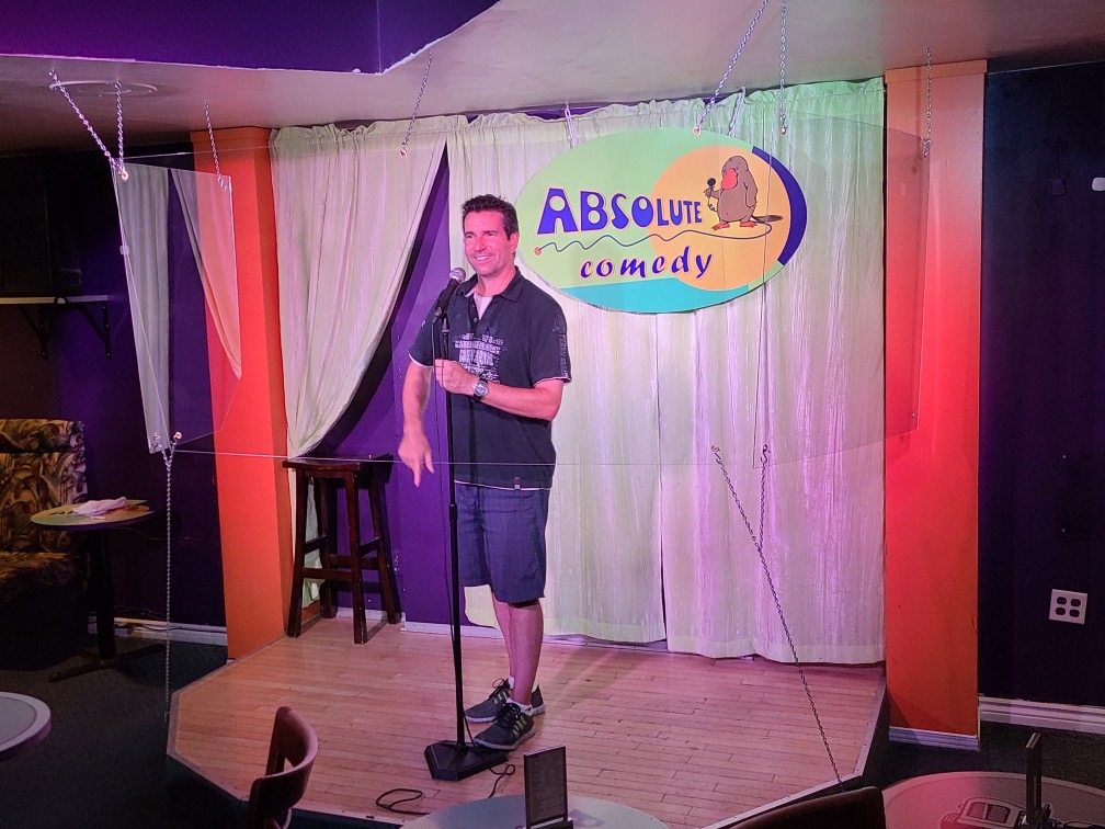 A photo of Jason Laurans, a comedian, performing behind plexiglass at Absolute Comedy.