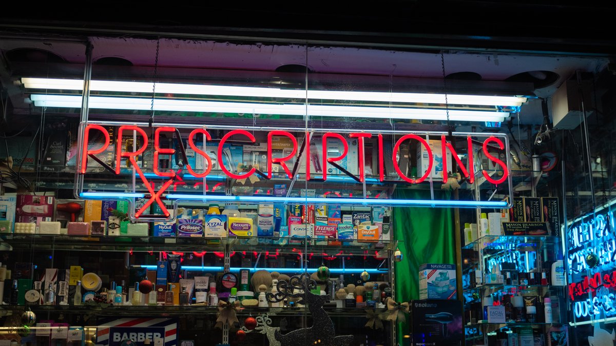 A red sign in a store reads, "Prescriptions." In the background, there are items and medication for sale.