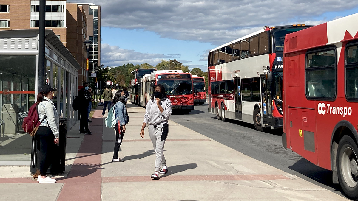 students wearing face coverings wait at the Carleton bus stop