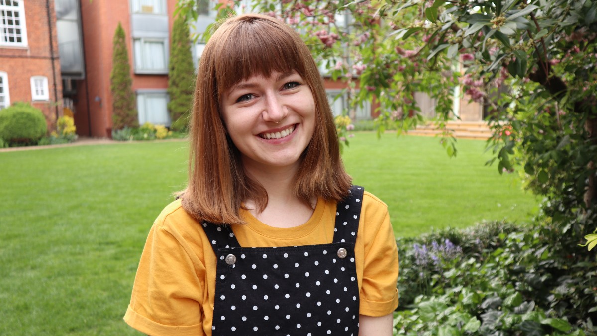 Chloe Halpenny tilts her head and smiles ooing into the camera. A courtyard and red building appear behind her. She is wearing a yellow shirt and overalls with black and white polkadots.