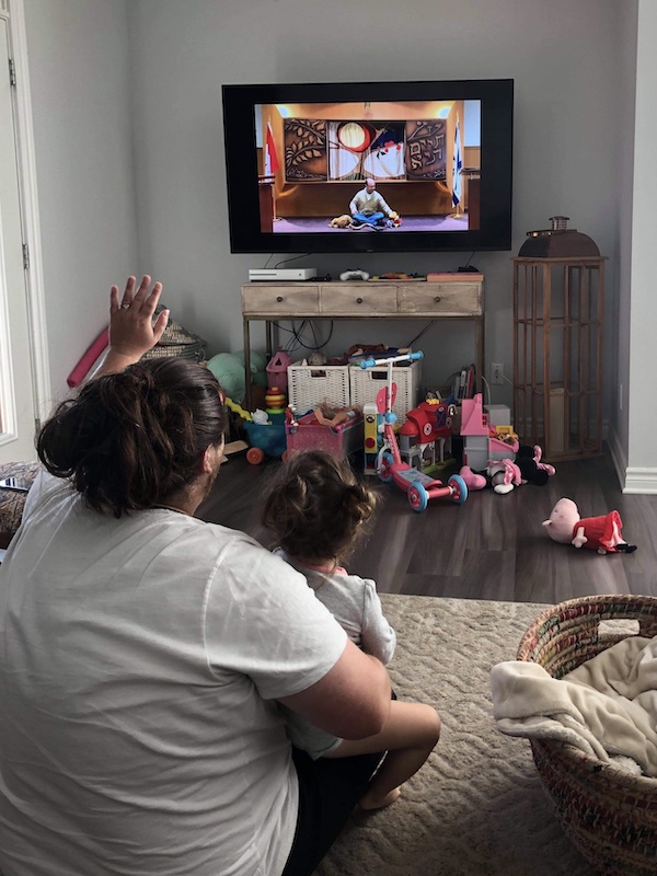 Dad sitting on the floor with daughter in his lap waving at the Television which is screening a Rosh Hashana service of a Rabbi sitting on the floor in front of a Shofar.