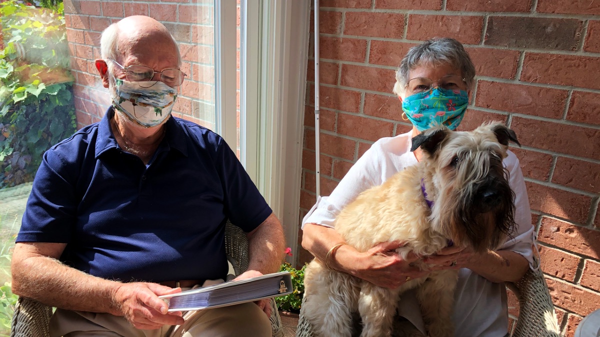 Two seniors, a man and a woman, sit on their patio. They are wearing masks. The man is looking at the cover of a photo album. The woman has a wheaten terrier on her lap.